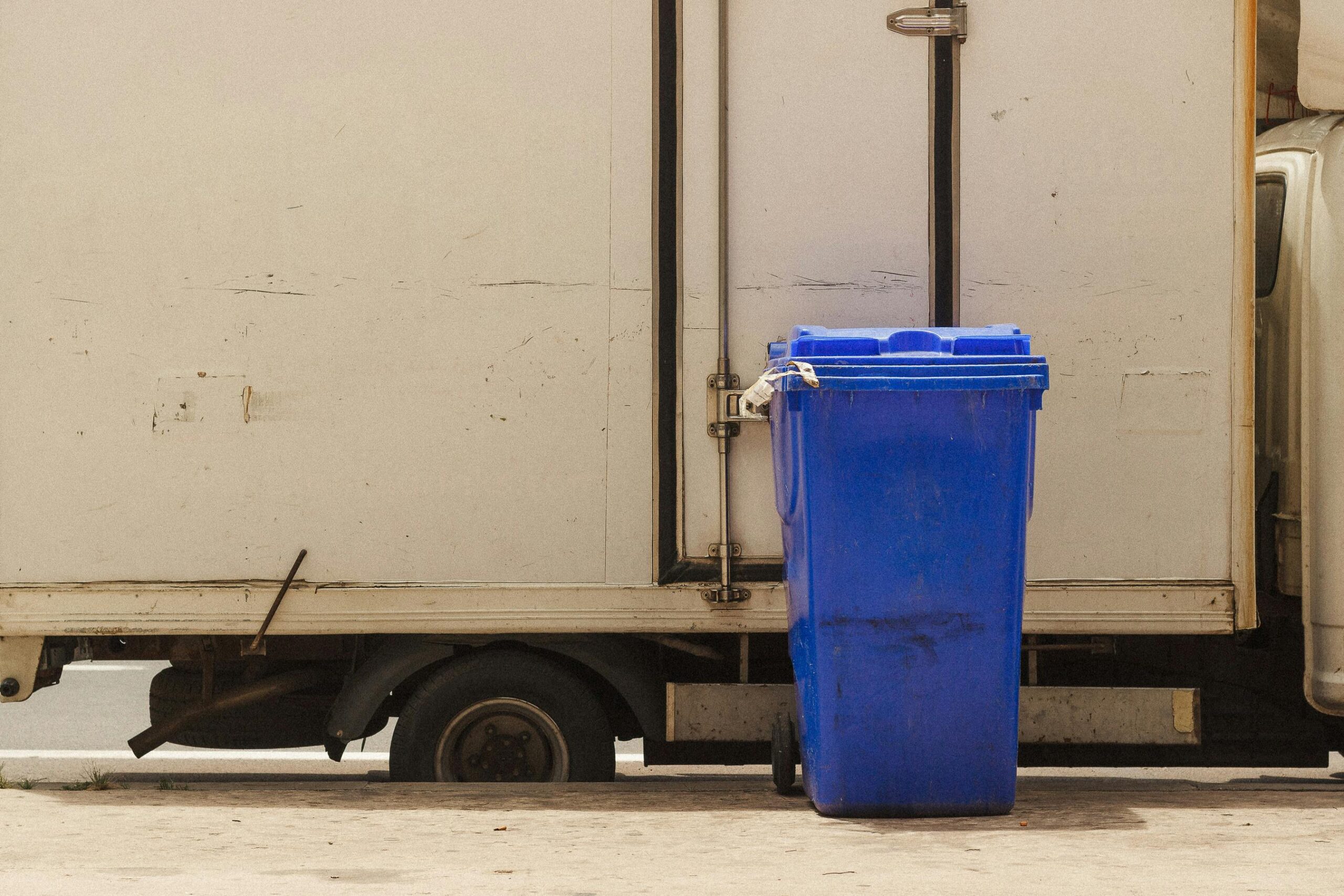 Blue recycling bin stands next to a parked delivery truck in an urban area.